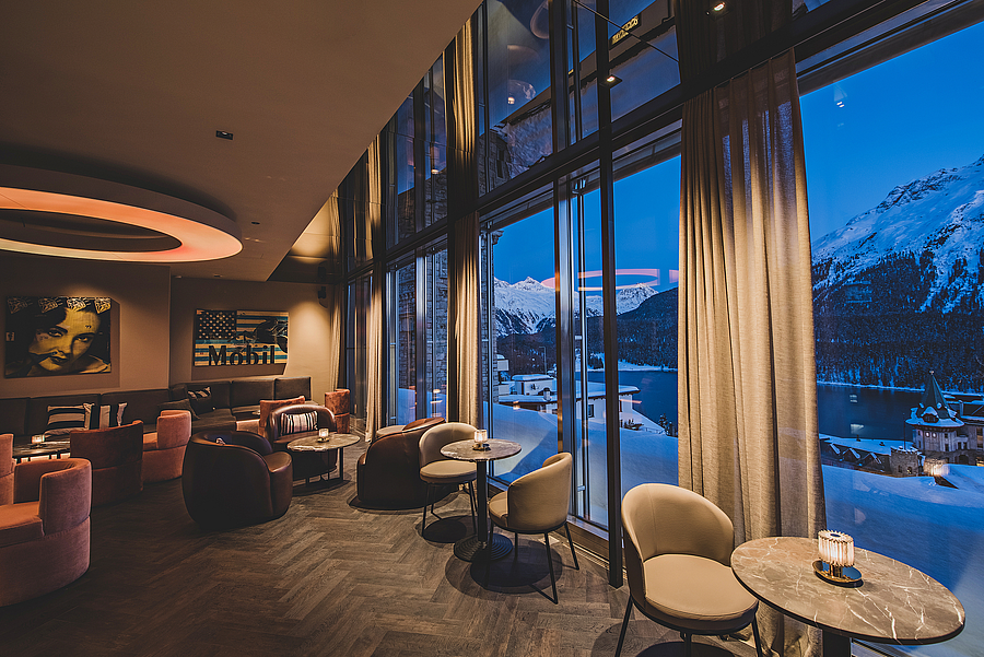 Elegante Bar mit großen Fenstern, die verschneite Berge und einen See in der Dämmerung zeigen.