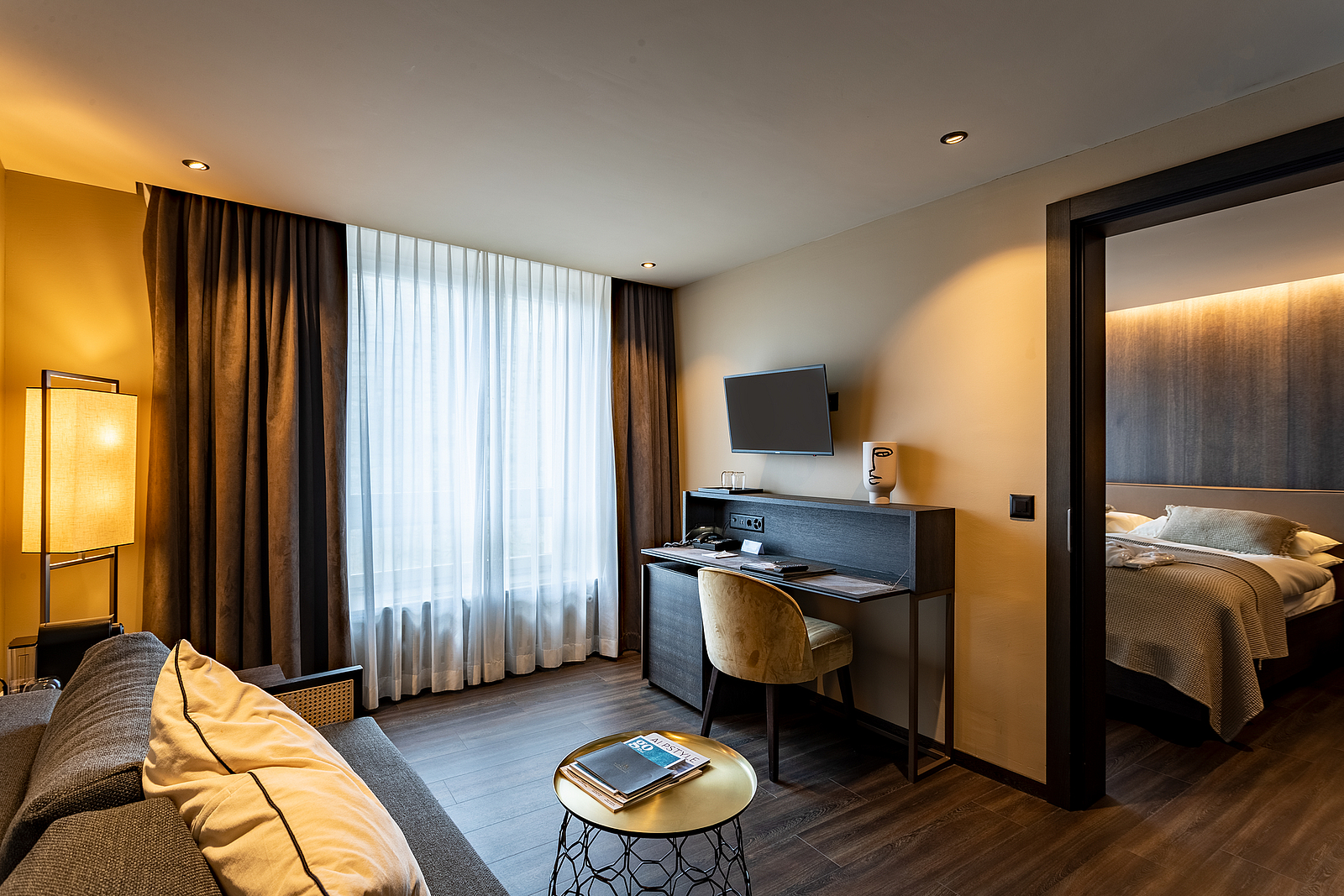 Modernes Hotelsuiten-Wohnzimmer mit Schreibtisch, TV, Sofa und Blick ins Schlafzimmer.