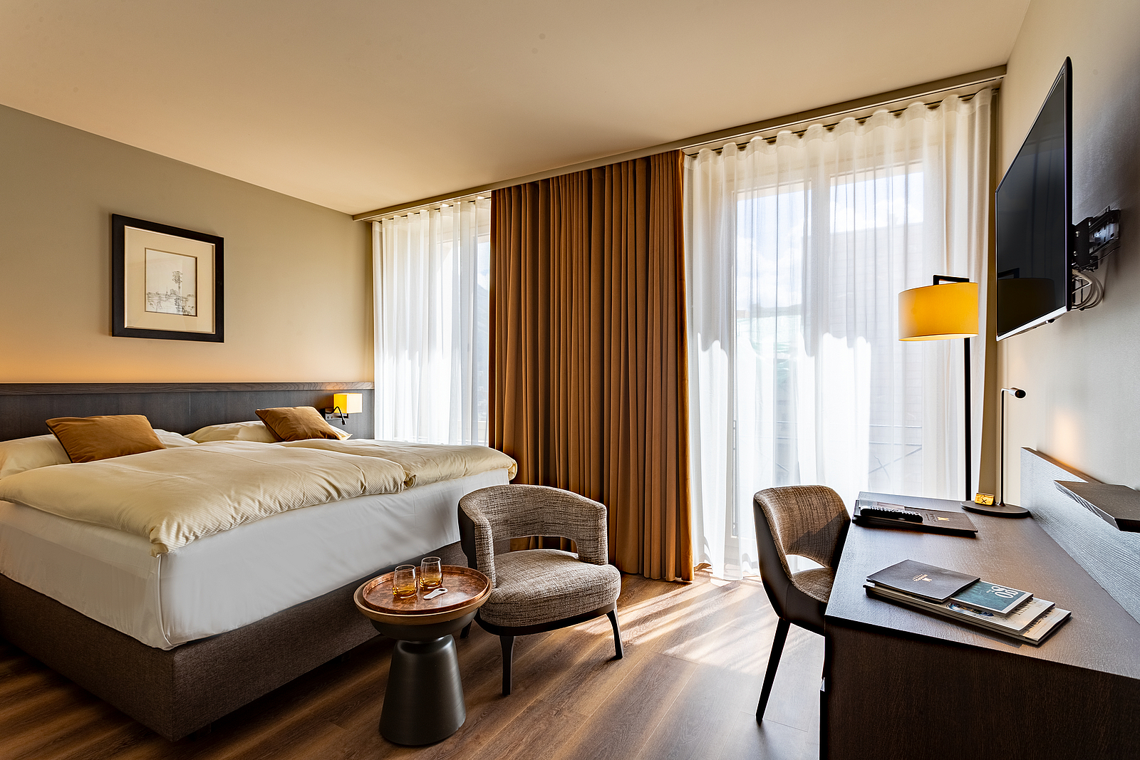 Modernes Hotelzimmer mit Bett, Schreibtisch, Sessel und Fenstern.