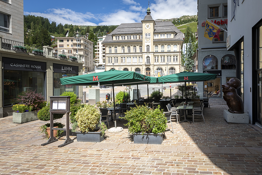 Rathausplatz in St. Moritz mit Außengastronomie unter grünen Schirmen.