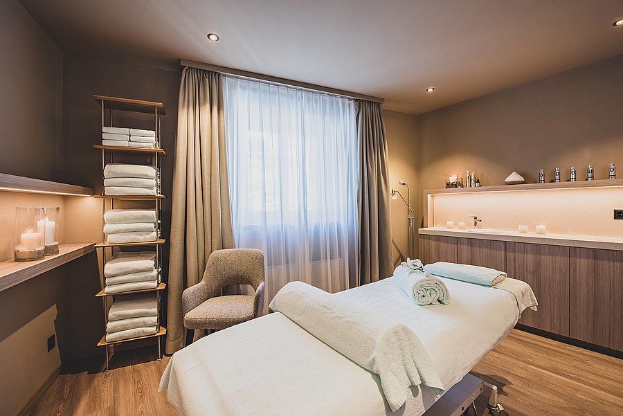 Ein ruhiger Spa-Raum mit Massageliege, Regalen voller Handtücher und sanfter Beleuchtung.