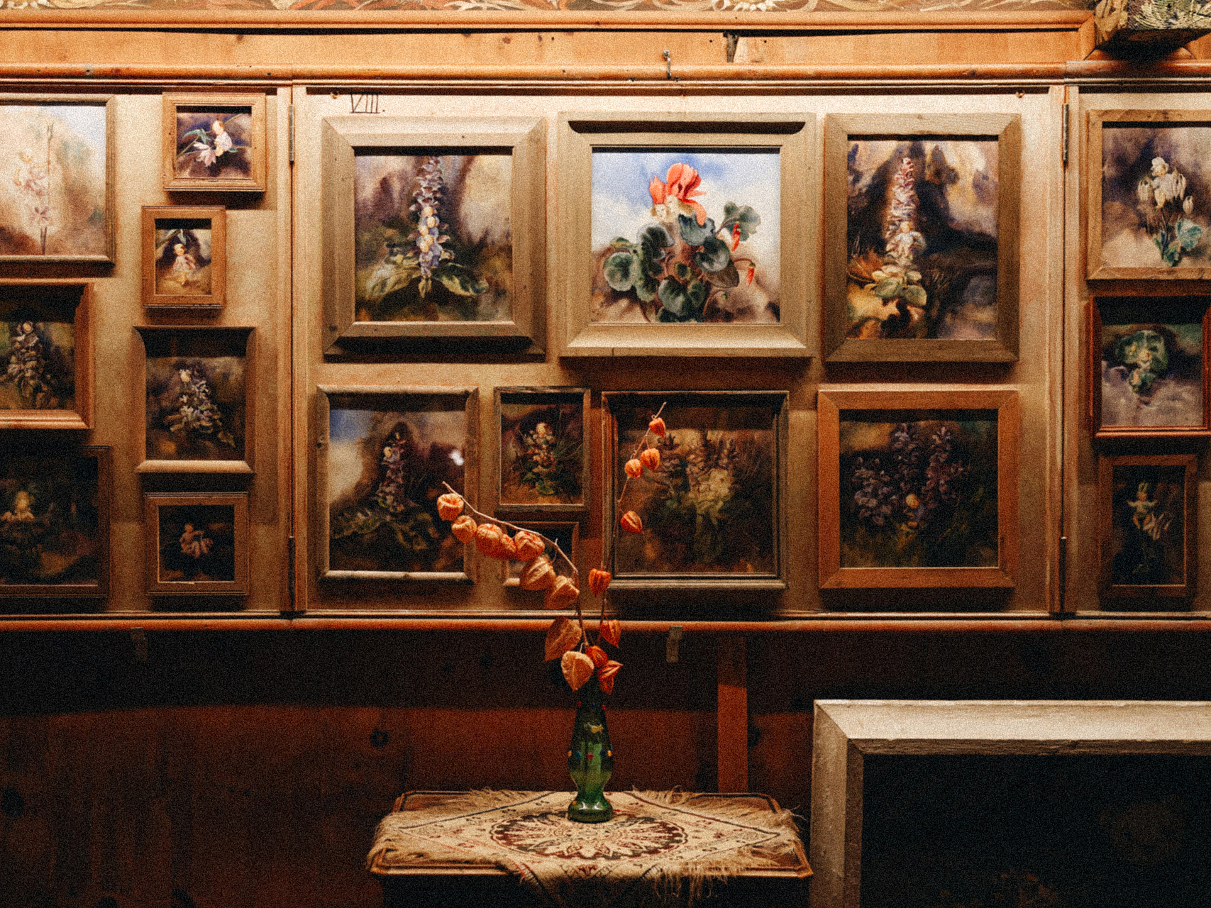 Galeriewand mit botanischen Gemälden Zahlreiche gerahmte Gemälde an einer Holzwand, davor eine Vase mit getrockneten Blumen auf einem Tisch.
