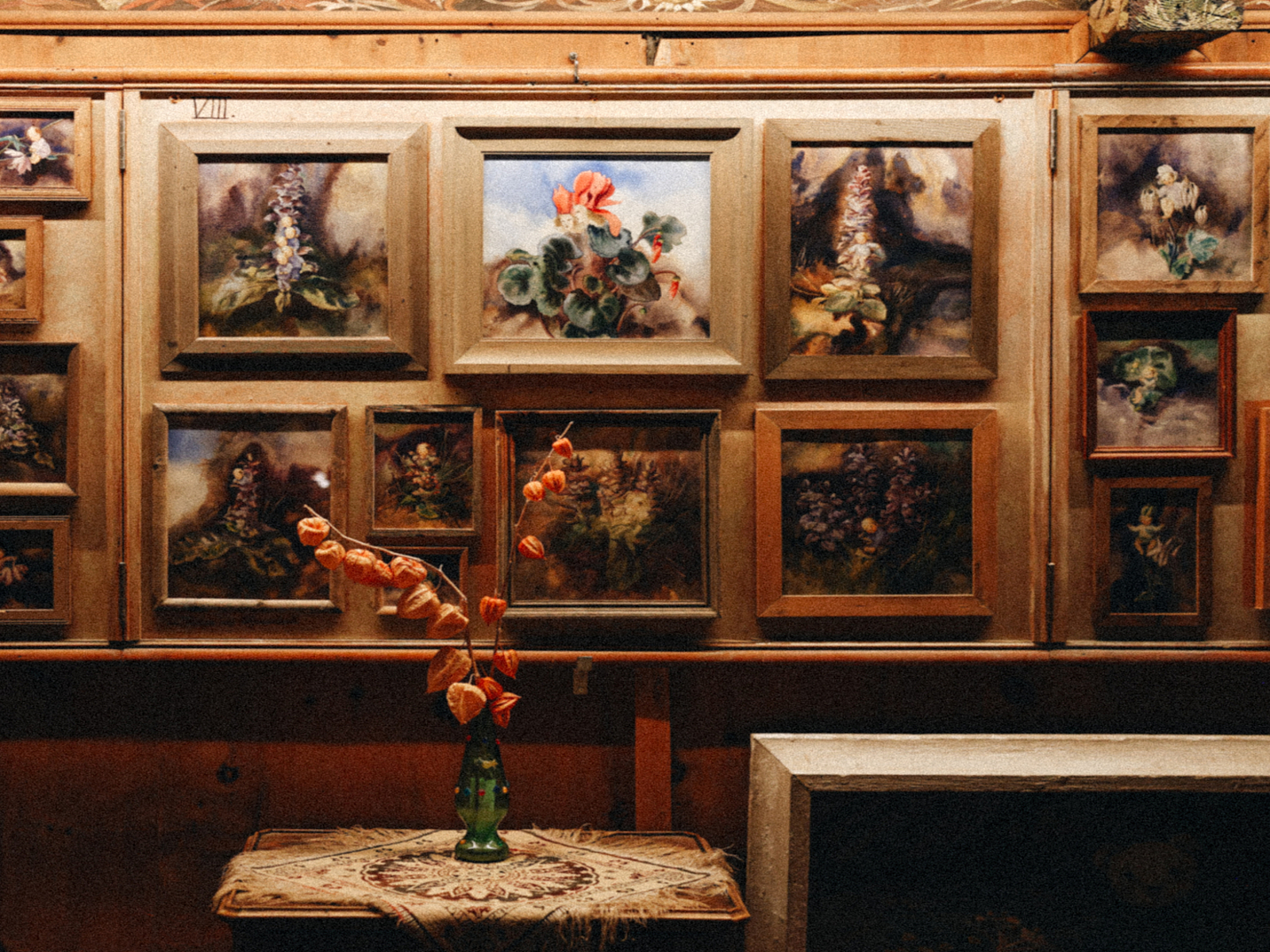 Galeriewand mit botanischen Gemälden Zahlreiche gerahmte Gemälde an einer Holzwand, davor eine Vase mit getrockneten Blumen auf einem Tisch.