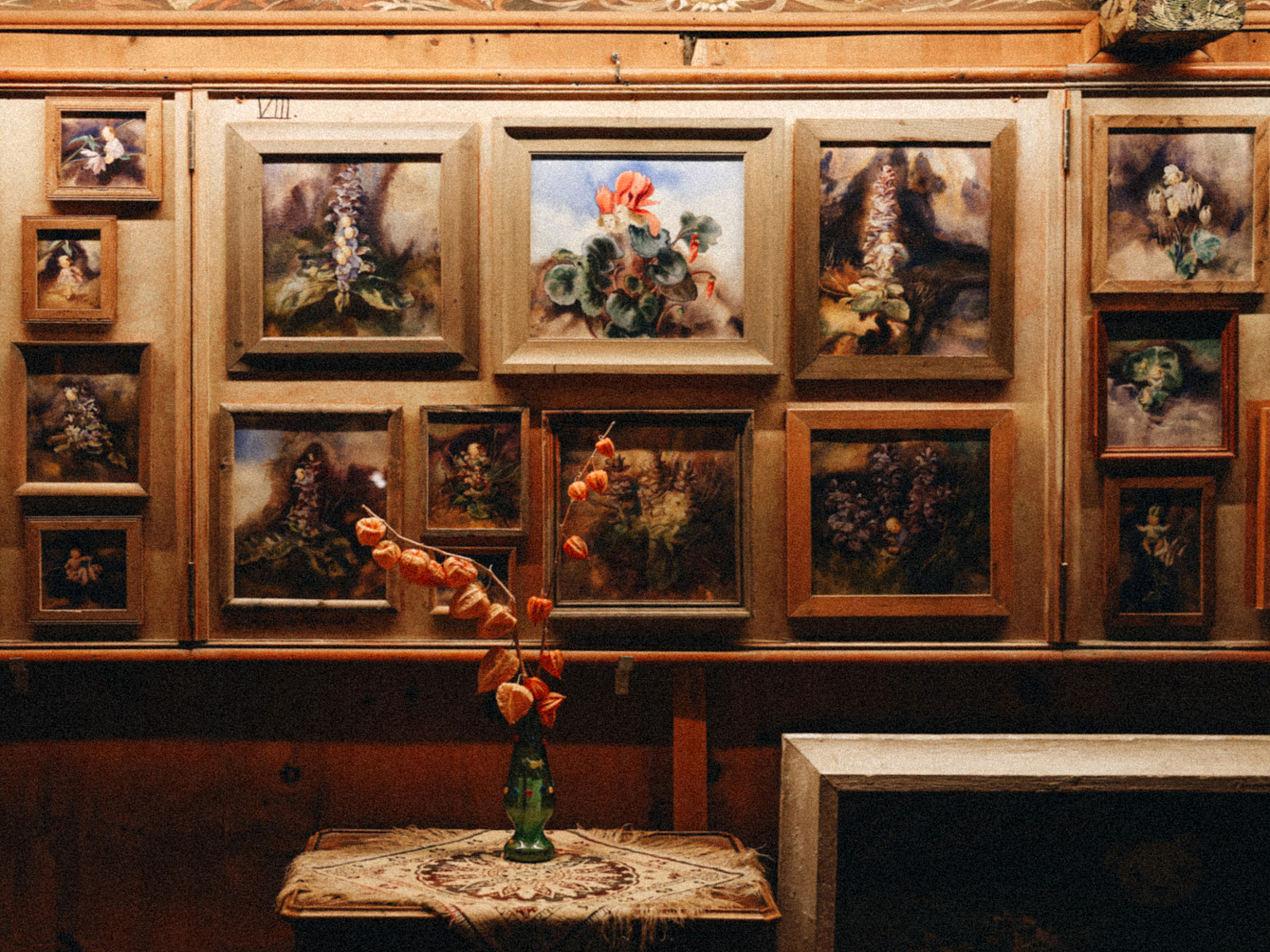 Galeriewand mit botanischen Gemälden Zahlreiche gerahmte Gemälde an einer Holzwand, davor eine Vase mit getrockneten Blumen auf einem Tisch.