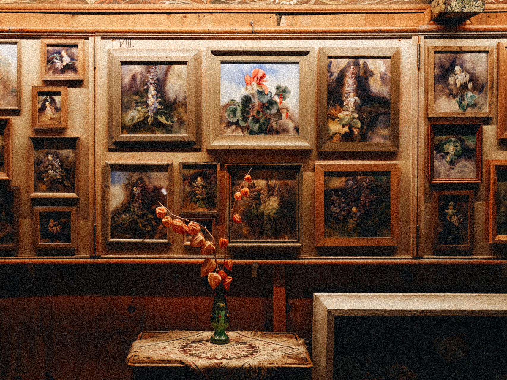 Galeriewand mit botanischen Gemälden Zahlreiche gerahmte Gemälde an einer Holzwand, davor eine Vase mit getrockneten Blumen auf einem Tisch.