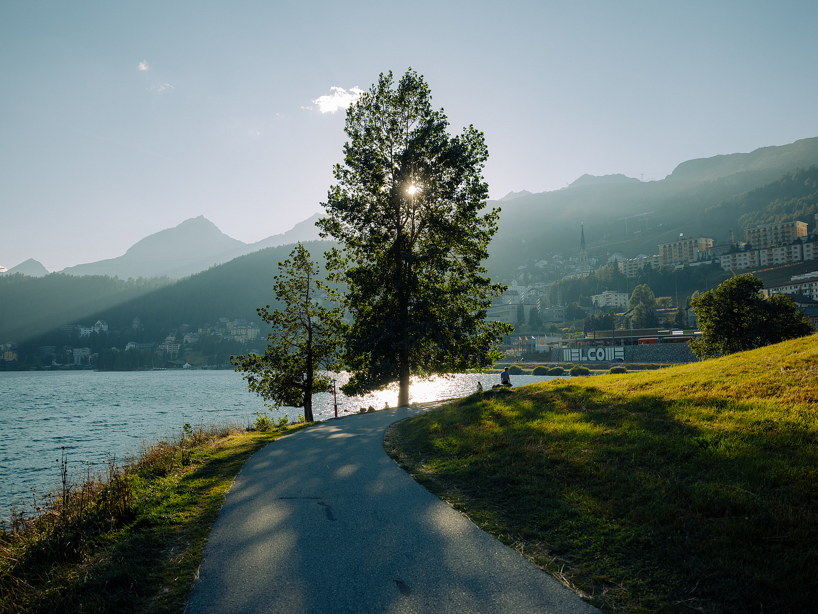 Ein malerischer Weg an einem See mit Bäumen und Bergen im Hintergrund.