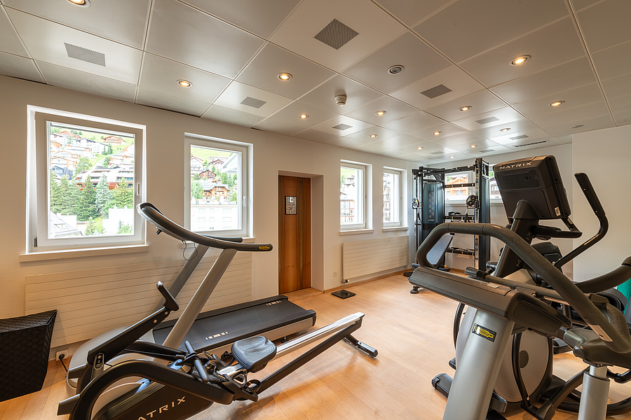 Helles Hotel-Fitnessstudio mit Laufband, Rudergerät, Heimtrainer und Fenstern mit Blick auf ein Bergdorf.