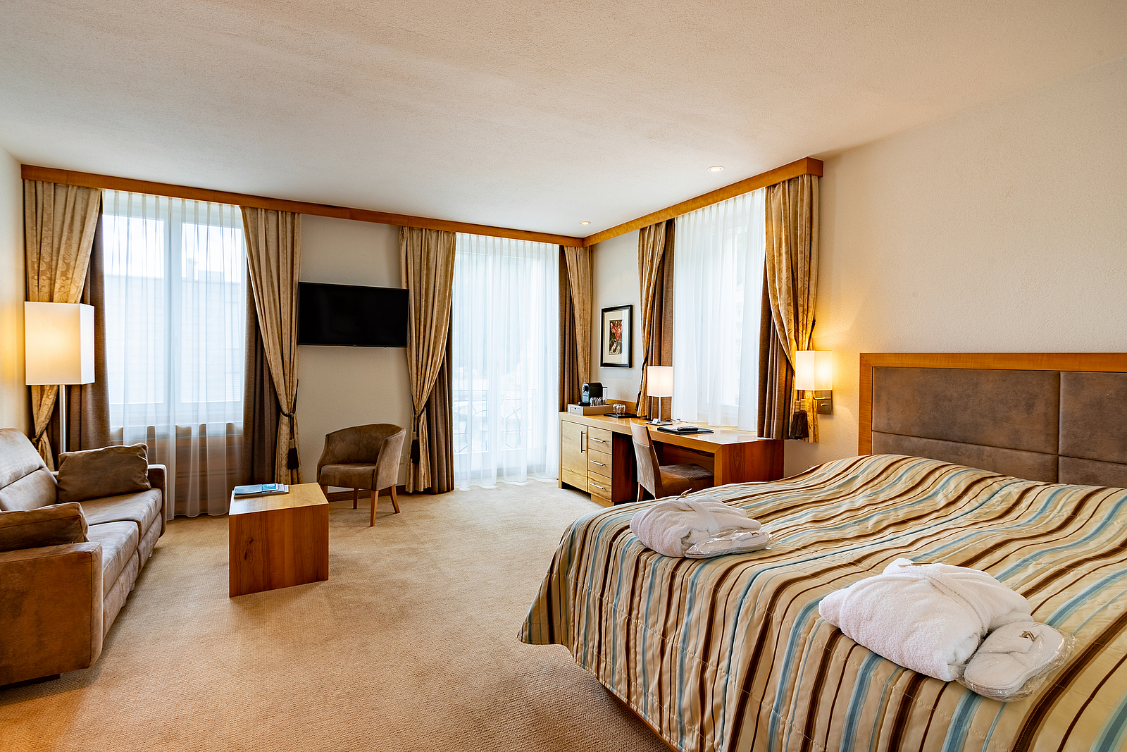 Geräumiges Hotelzimmer mit großem Bett, Sitzecke, Schreibtisch und großen Fenstern.