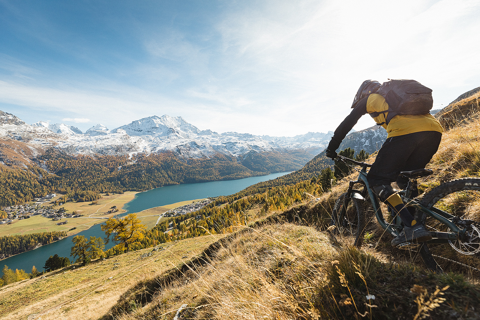 Ein Mountainbiker auf einem steilen Alpinen Trail mit Blick auf See und schneebedeckte Berge.