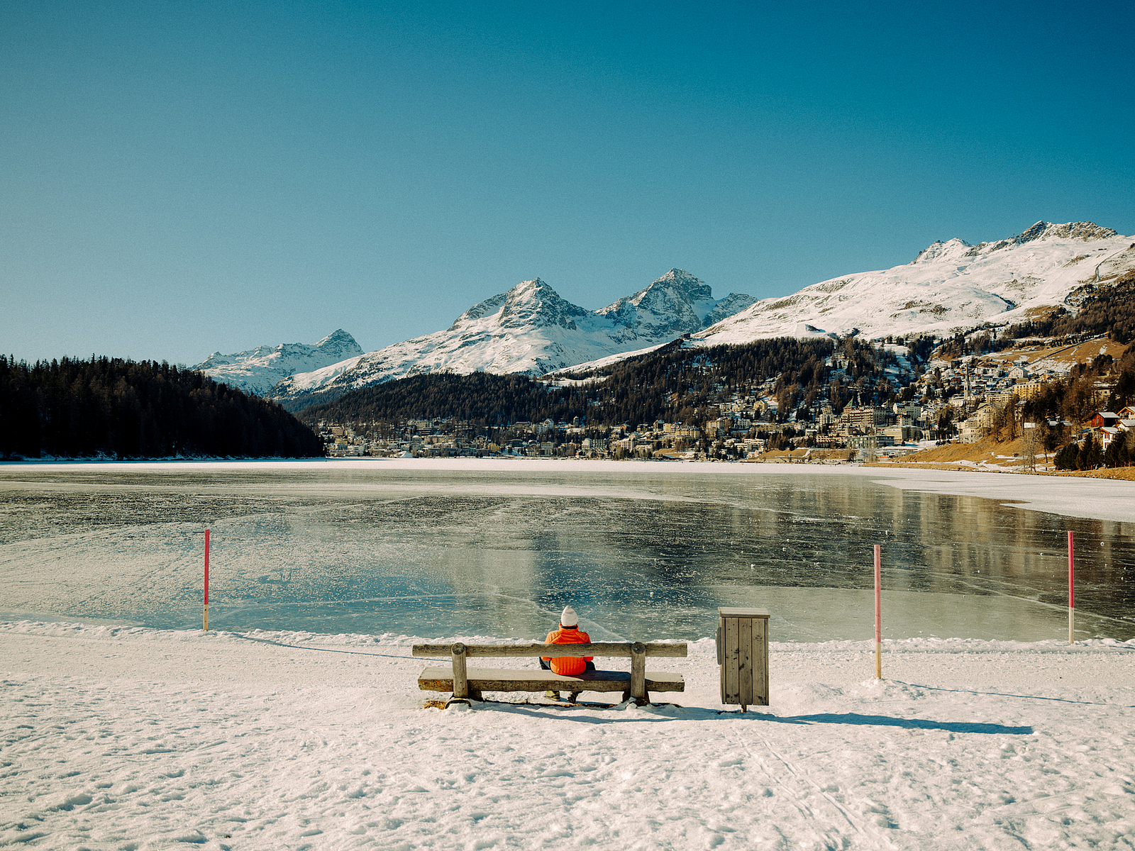 Person auf Bank am gefrorenen See Person in orangefarbener Jacke sitzt auf Bank vor gefrorenem See und Bergen.