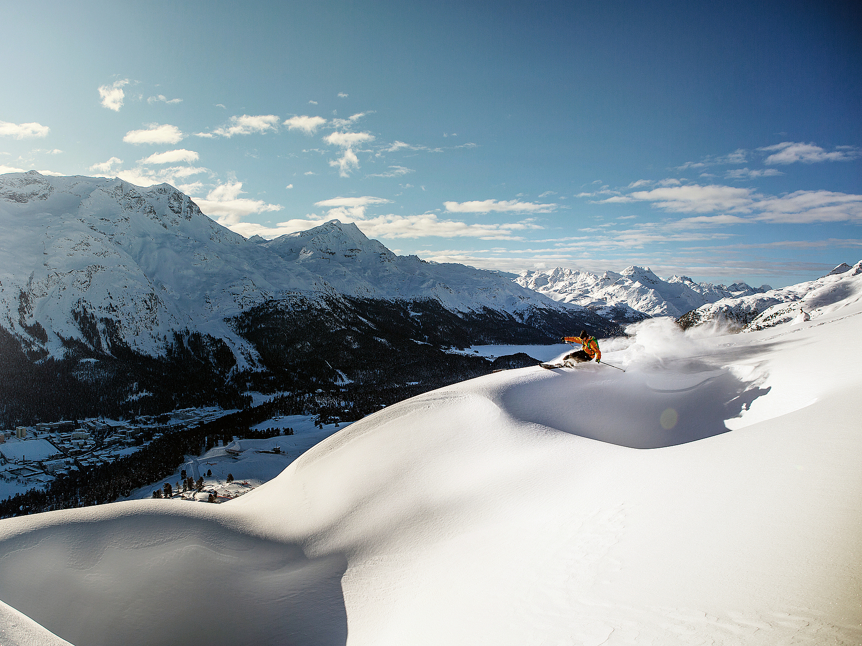 Skifahren in den Schweizer Alpen Skifahrer in gelber Jacke wirbelt Pulverschnee auf einer Bergkratte mit Alpenpanorama auf.