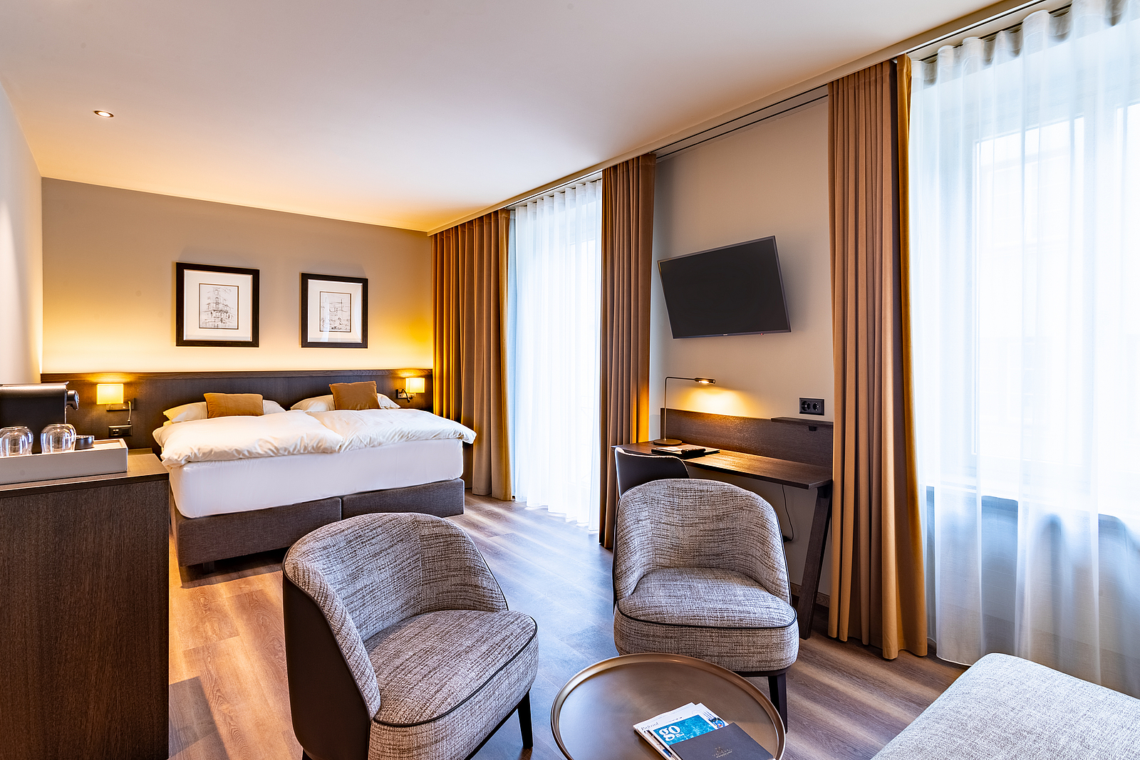 Modernes Hotelzimmer mit Bett, Sesseln, Schreibtisch und großen Fenstern.
